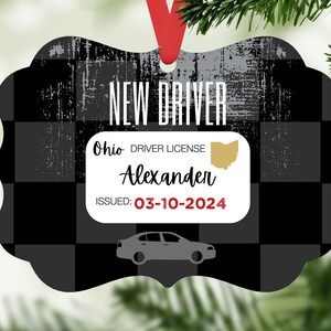 Może przedstawiać: Ozdoba dekoracyjna z czarno-szarym wzorem w kratę i napisem "NEW DRIVER". Zawiera fałszywe prawo jazdy z Ohio z imieniem "Alexander" i datą wydania "03-10-2024". Czerwona wstążka jest przymocowana do zawieszenia.