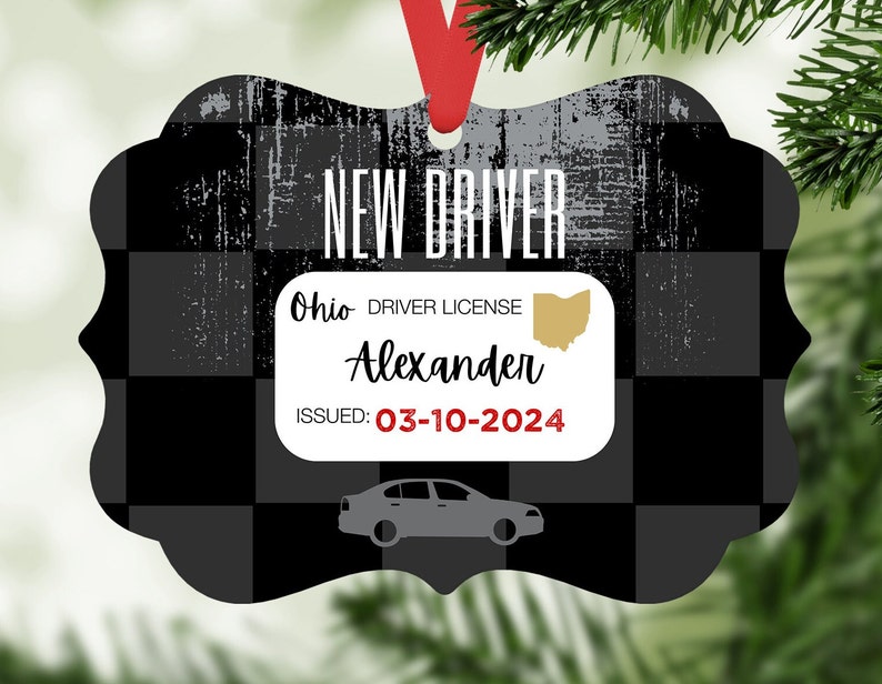 Może przedstawiać: Ozdoba dekoracyjna z czarno-szarym wzorem w kratę i napisem "NEW DRIVER". Zawiera fałszywe prawo jazdy z Ohio z imieniem "Alexander" i datą wydania "03-10-2024". Czerwona wstążka jest przymocowana do zawieszenia.