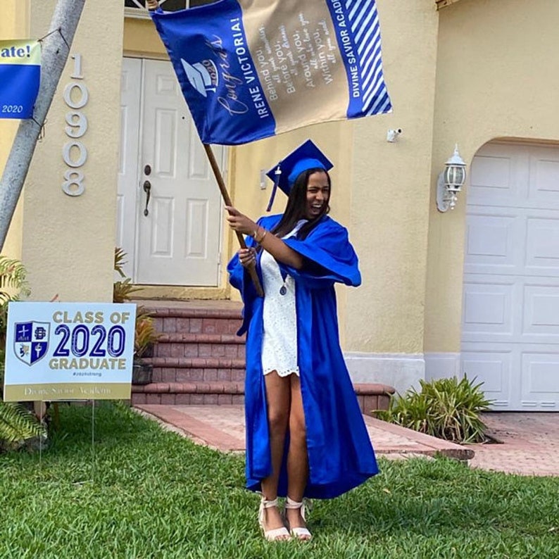 May include: A graduate in a blue cap and gown holds a flag that reads "Congras Irene Victoria!" and "Divine Savior Academy." A sign in the yard says "Class of 2020 Graduate." The graduate is wearing a white dress and pink heels.