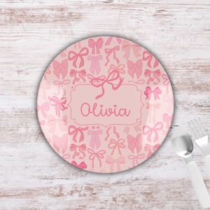 May include: A pink plate with a pattern of bows in varying shades of pink. The plate has a light pink background and features the name "Olivia" in a decorative frame. A spoon and fork are visible.