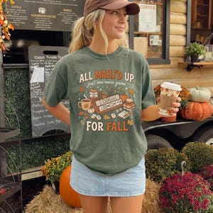 May include: A sage green short-sleeve t-shirt featuring a graphic print with the text "ALL MAHJ'D UP just one more game FOR FALL". The design includes illustrations of mahjong tiles, a coffee mug, a pumpkin, and autumn leaves.