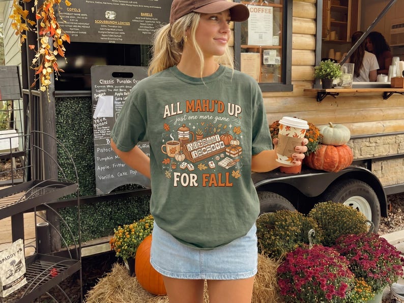 May include: A sage green short-sleeve t-shirt featuring a graphic print with the text "ALL MAHJ'D UP just one more game FOR FALL". The design includes illustrations of mahjong tiles, a coffee mug, a pumpkin, and autumn leaves.