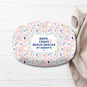 May include: An oval serving platter with a pink background and a pattern of playing cards featuring seahorses, octopuses, and other sea creatures. The platter has the text "BAMS, CRAKS & BEACH SNACKS at Sarah's".