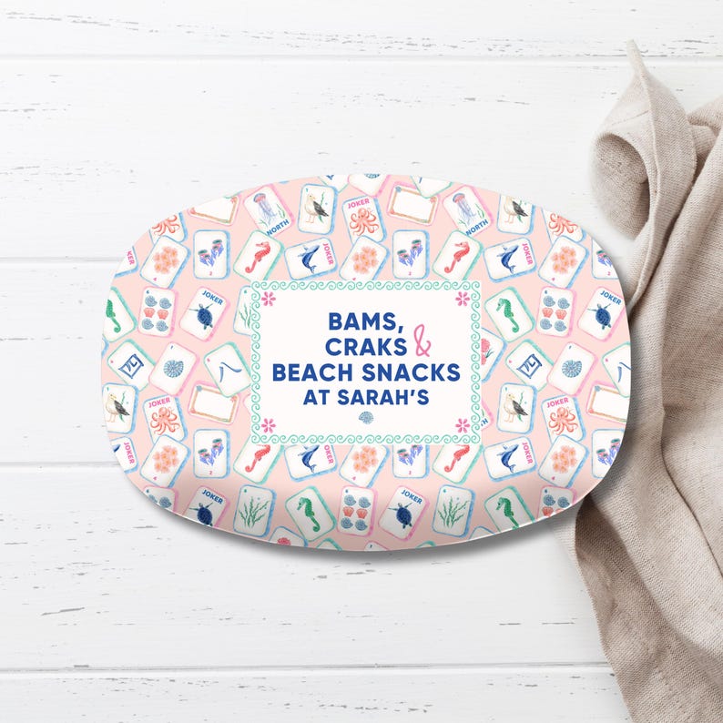 May include: An oval serving platter with a pink background and a pattern of playing cards featuring seahorses, octopuses, and other sea creatures. The platter has the text "BAMS, CRAKS & BEACH SNACKS at Sarah's".
