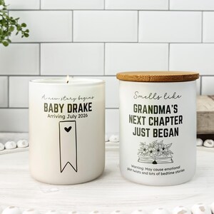 Może przedstawiać: Dwa białe świece z czarnym tekstem. Na jednej napisano "Baby Drake Arriving July 2026", a na drugiej "Grandma's Next Chapter Just Began". Świeca po prawej stronie ma drewnianą pokrywkę. Świece stoją na białej tacy.
