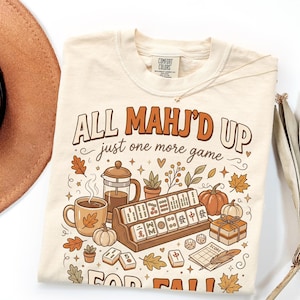 May include: A cream-colored t-shirt featuring a graphic print with the text "ALL MAHJ'D UP just one more game FOR FALL". The illustration includes a coffee mug, a French press, pumpkins, autumn leaves, and a wooden rack filled with mahjong tiles.