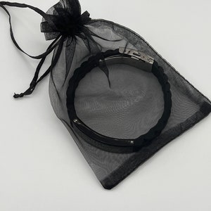 Op de afbeelding: Een zwarte armband met een gevlochten ontwerp en een zilveren sluiting wordt gepresenteerd in een doorzichtige zwarte trekkoordtas. De armband lijkt gemaakt te zijn van een flexibel materiaal.
