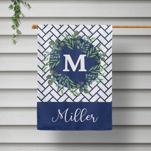 May include: A navy blue garden flag with a white and navy blue geometric pattern. A white letter "M" is surrounded by a wreath of green leaves on a navy blue circle. The text "Miller" is printed in white cursive script on the bottom of the flag.