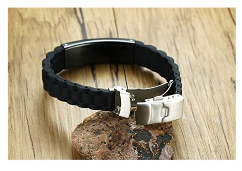 Op de afbeelding: Een zwarte siliconen armband met een roestvrijstalen sluiting. De armband heeft een getextureerd, geribbeld ontwerp en wordt open getoond, waarbij het sluitmechanisme zichtbaar is. De armband ligt op een kleine, getextureerde steen tegen een houten achtergrond.
