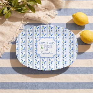 May include: An oval serving platter with a blue and white striped pattern and a repeating design of blue berries and green leaves. The platter has the text "BAMS, CRAKS & SNACKS AT Sarah's" in a decorative frame. Two lemons are on the striped surface.