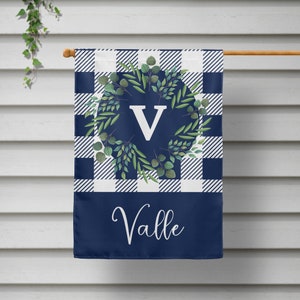 May include: A navy blue and white checkered garden flag with a green wreath and the letter "V" in the center. The bottom of the flag is solid navy blue with the name "Valle" in white cursive writing.