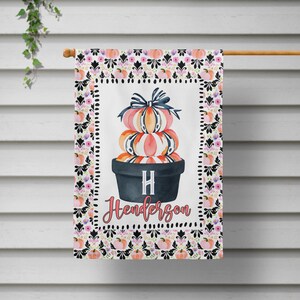 May include: A decorative garden flag featuring a stack of pumpkins in a black pot, topped with a blue bow. The flag has a white background with the name "Henderson" and the initial "H". The border has a floral pattern.