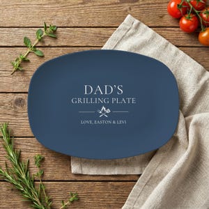 May include: A navy blue oval grilling plate with white text that reads "DAD'S GRILLING PLATE" and "LOVE, EASTON & LEVI". The plate has a graphic of a flame and crossed grilling tools. The plate is on a wooden surface.