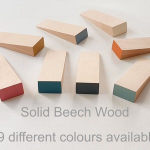 May include: Nine wooden doorstops in various colours, including red, orange, teal, and grey. The doorstops are made of solid beech wood and are all the same size and shape. The text "Solid Beech Wood" and "9 different colours available" is printed below the doorstops.