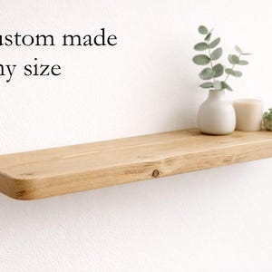 May include: A natural wood floating shelf with rounded corners, fixed to a white wall. The shelf displays a small white vase with foliage, a candle, and a small potted plant. Text on the wall reads "Custom made Any size."