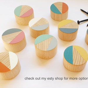 May include: Nine wooden wall hooks with geometric designs in shades of teal, yellow, pink, and white. The hooks are round and have a natural wood finish.