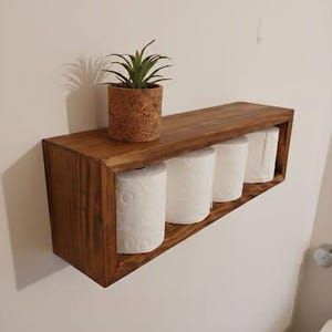 May include: A stained wooden bathroom shelf holding four white toilet paper rolls. A small potted plant sits on top. The shelf is mounted on a white wall.