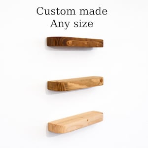 May include: Three wooden floating shelves in shades of brown, mounted on a white wall. The top shelf is a dark brown, the middle shelf is a lighter brown, and the bottom shelf is natural wood. Text above reads "Custom made Any size".