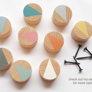 May include: Eight wooden wall hooks with a geometric triangle design in various colours, including teal, light blue, pink, orange, yellow, and grey. The hooks are displayed on a white surface with black screws.