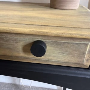 May include: A wooden drawer with a black knob. The drawer is painted in a light brown colour with a distressed finish. The drawer is part of a black painted table.