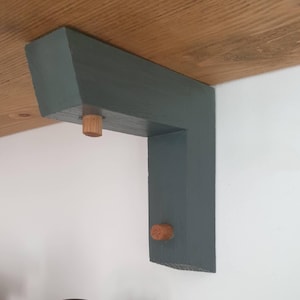 May include: A dark teal L-shaped wooden shelf bracket with a natural wood dowel pin. The bracket is mounted to a light brown wooden shelf. The wall is white. The bracket is designed to support a shelf.