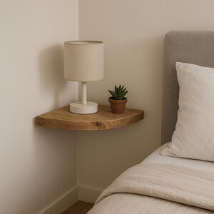 May include: A wooden corner shelf with a small white lamp and a potted succulent. The lamp has a beige lampshade. The shelf is mounted on a white wall next to a bed with a white pillow. The shelf is made of wood.