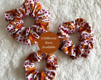 May include: Three gingerbread man patterned scrunchies in different sizes. The scrunchies are white with brown gingerbread men and pink polka dots. The text "3 Different Sizes Available" is written on a brown circle.