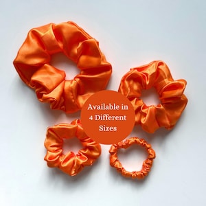 May include: Four orange satin scrunchies in different sizes. The scrunchies are displayed on a white background. A circular graphic in the centre reads "Available in 4 Different Sizes".