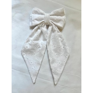 May include: A white hair bow made of eyelet fabric featuring a delicate floral embroidery pattern with small circular cutouts. The bow has a central knot and two long, pointed tails that hang downwards, showcasing the intricate needlework details.