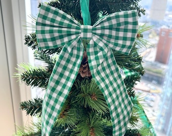 May include: A green and white gingham bow adorns a small artificial Christmas tree. The bow is made of fabric and has long, flowing tails. A small pinecone is nestled beneath the bow. Green ribbon is used to hang the bow on the tree.