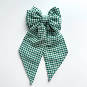 May include: A large green and white gingham bow, likely for hair or as a decorative accessory. The bow features a classic checkered pattern and long, pointed tails. The fabric appears to be a lightweight material.