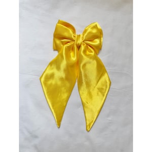May include: A bright yellow hair bow crafted from a smooth, lustrous satin fabric. The accessory features a central knot with two large, voluminous loops at the top and two long, pointed tails extending downward, creating a classic bow shape.