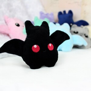 May include: A black plush bat with red eyes and large wings is in focus, with other colorful plush bats blurred in the background. The plush toys are in various colors, including pink, blue, and gray. The bats have a cute, cartoonish design.
