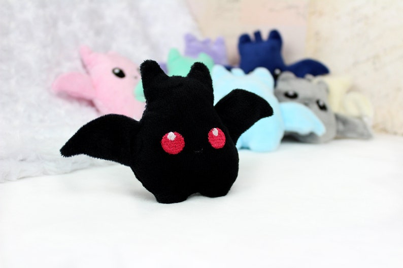 May include: A black plush bat with red eyes and large wings is in focus, with other colorful plush bats blurred in the background. The plush toys are in various colors, including pink, blue, and gray. The bats have a cute, cartoonish design.