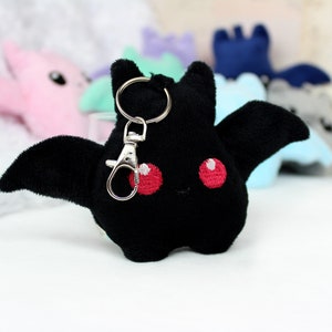May include: A black plush bat keychain with red embroidered eyes and a silver clasp. The bat has outstretched wings and is part of a collection of plush bats in various pastel colors.
