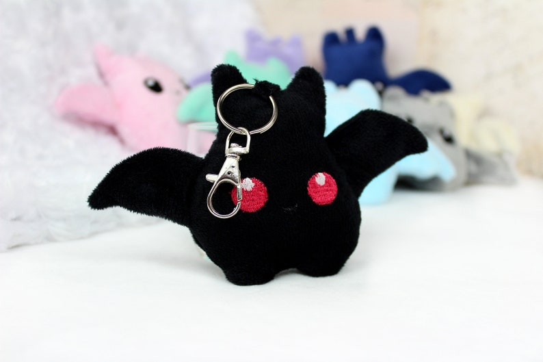 May include: A black plush bat keychain with red embroidered eyes and a silver clasp. The bat has outstretched wings and is part of a collection of plush bats in various pastel colors.