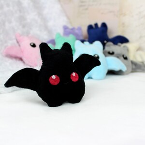 May include: A black plush bat with red eyes and outstretched wings is in focus, with several other colorful plush bats blurred in the background. The bats are in various pastel colors, including pink, blue, and gray. The plush toys are on a white surface.