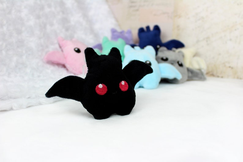 May include: A black plush bat with red eyes and outstretched wings is in focus, with several other colorful plush bats blurred in the background. The bats are in various pastel colors, including pink, blue, and gray. The plush toys are on a white surface.