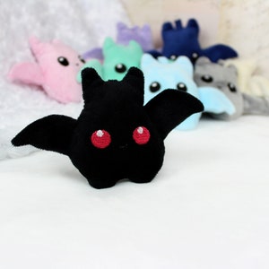 May include: A black plush bat with red eyes and outstretched wings is the focal point, surrounded by other colorful plush bats in pink, blue, green, and gray. The toys are on a white surface, creating a playful and whimsical scene.
