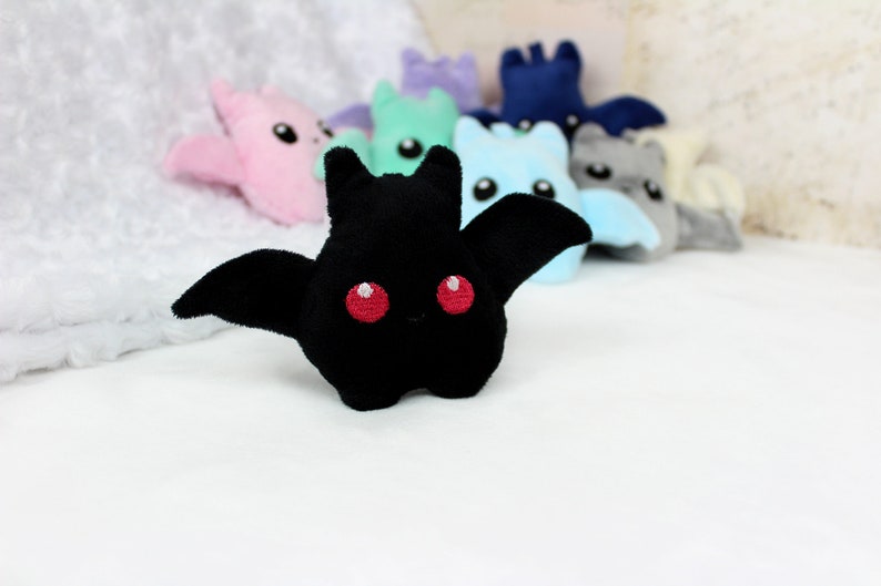 May include: A black plush bat with red eyes and outstretched wings is the focal point, surrounded by other colorful plush bats in pink, blue, green, and gray. The toys are on a white surface, creating a playful and whimsical scene.