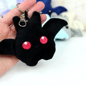 May include: Black plush bat keychain with embroidered red eyes. The bat has outstretched wings and a small smile. Other plush bats in various colors are visible in the background.