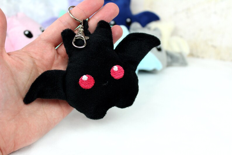 May include: Black plush bat keychain with embroidered red eyes. The bat has outstretched wings and a small smile. Other plush bats in various colors are visible in the background.