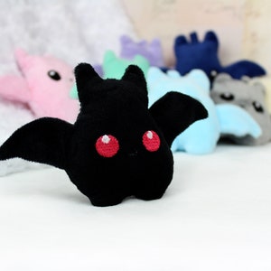 May include: A plush black bat with red embroidered eyes and wings is in focus, with other colorful bat plushies blurred in the background. The plush toys are in various pastel colors, including pink, blue, and gray.