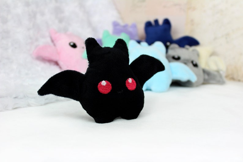 May include: A plush black bat with red embroidered eyes and wings is in focus, with other colorful bat plushies blurred in the background. The plush toys are in various pastel colors, including pink, blue, and gray.
