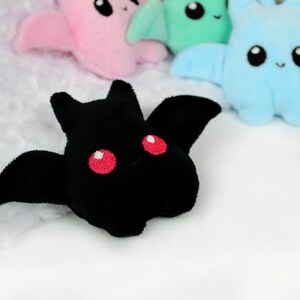 May include: A black plush bat with red embroidered eyes and wings is in focus, with other colorful plush bats in the background. The bats have a cute, cartoonish design, with embroidered faces and wings. The background is a soft, white, textured fabric.