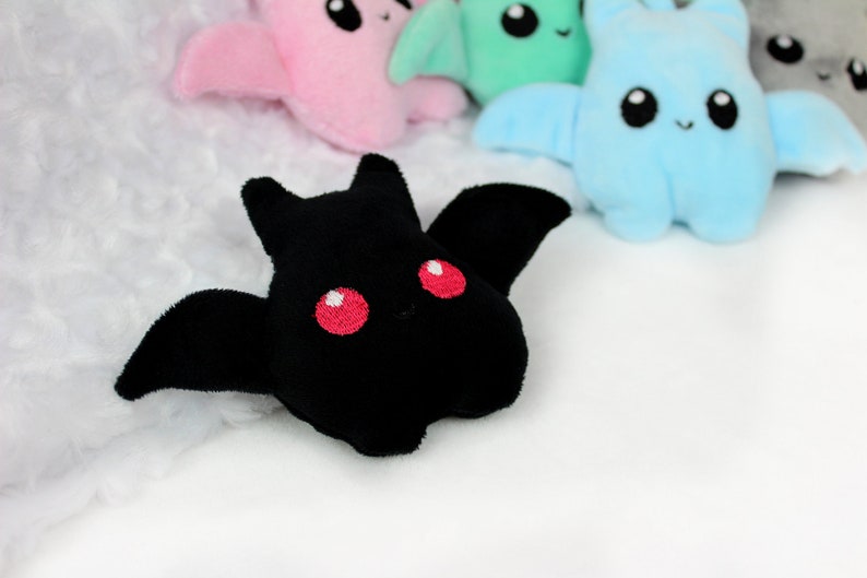 May include: A black plush bat with red embroidered eyes and wings is in focus, with other colorful plush bats in the background. The bats have a cute, cartoonish design, with embroidered faces and wings. The background is a soft, white, textured fabric.