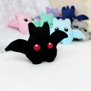May include: A black plush bat with red embroidered eyes and outstretched wings is in focus, with several other colorful plush bats blurred in the background. The bats are in shades of pink, green, blue, and gray.
