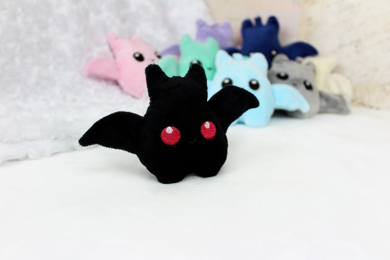 May include: A black plush bat with red embroidered eyes and outstretched wings is in focus, with several other colorful plush bats blurred in the background. The bats are in shades of pink, green, blue, and gray.