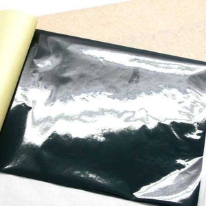 May include: A sheet of black, glossy material with a reflective surface, partially peeled back to reveal a yellow adhesive backing. A tan paper is visible in the background.