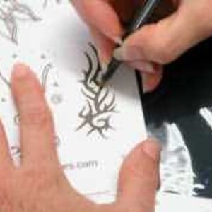May include: A hand holding a black pen is drawing a tribal design on white paper. The design is a series of curved lines and swirls. Other partially drawn designs are visible on the paper. The website address is visible at the bottom of the page.